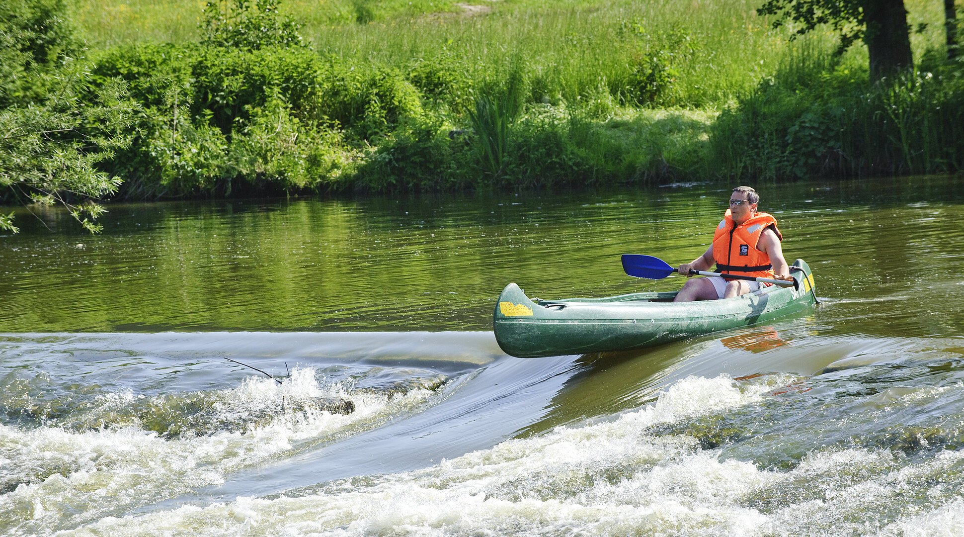 Ein Bootsfahrer mit oranger Rettungweste und Paddel steht mit seinem grünen Kanu am Anfang der Bootstreppe in der Altmühl. Das Wasser rauscht hier an dem steinernen Wehr hinunter. Die Bootsrutsche ist eine Vertiefung für die Fische und auch Boote. Das Ufer ist sattgrün auf dem die Sonne scheint.