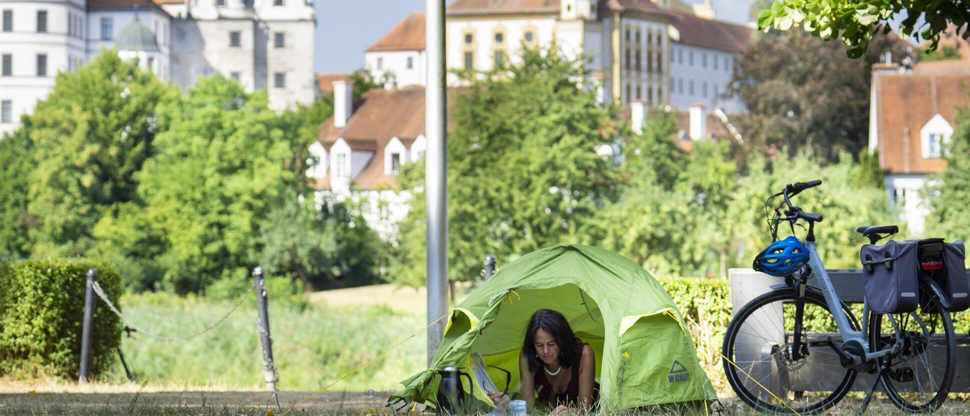 Eine Frau in einem grünen Ein-Mann-Zelt schaut heraus. Rechts neben ihr steht ein Fahrrad. Das Schloss thront hinter ihr.