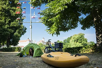 Ein Kajak und ein grünes Zelt sind auf dem Bootsübernachtungsplatz. Im Hintergrund grüne Bäume, ein Fahrrad und hellblauer Himmel.