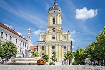 Radelnde auf dem Karlsplatz vor der prächtigen Hofkirche. Im Vordergrund ist ein Brunnen zu sehen.