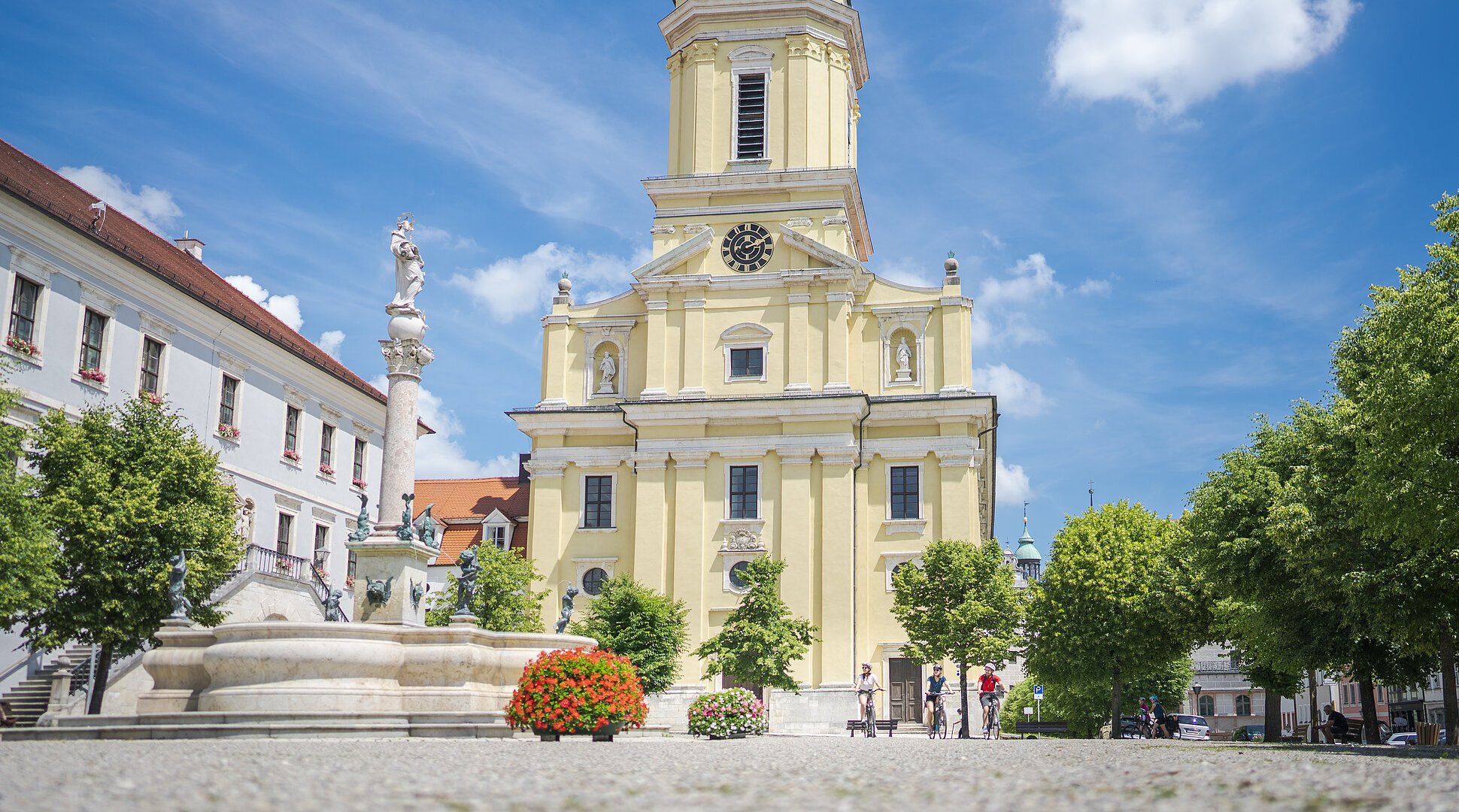 Radelnde auf dem Karlsplatz vor der prächtigen Hofkirche. Im Vordergrund ist ein Brunnen zu sehen.