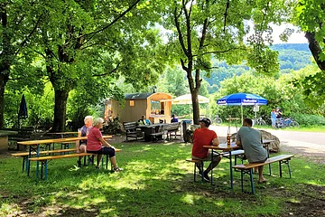 Blick auf den Schäferwagen und Rastplatz "Mein Platz an der Bruck". Im Vordergrund Bierbänke mit Gästen, im Hintergrund sieht man den umgebauten Schäferwagen und ankommende Radfahrer.