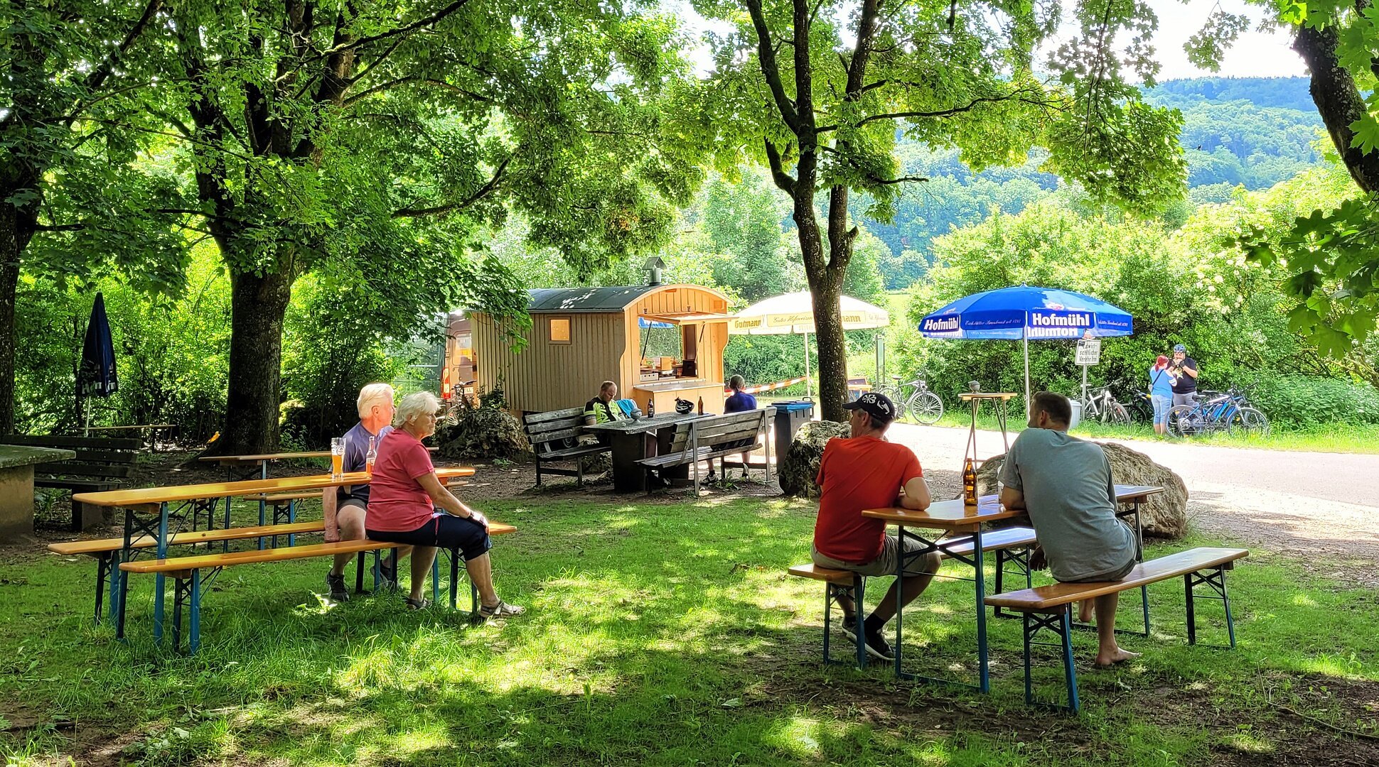 Blick auf den Schäferwagen und Rastplatz "Mein Platz an der Bruck". Im Vordergrund Bierbänke mit Gästen, im Hintergrund sieht man den umgebauten Schäferwagen und ankommende Radfahrer.
