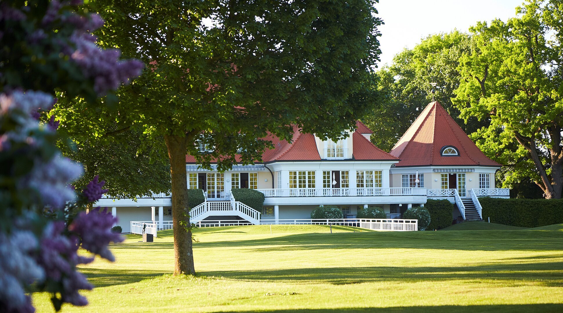 Ein großes Haus mit roten Dächern und weißen Fensterrahmen, umgeben von Bäumen und Rasenfläche.