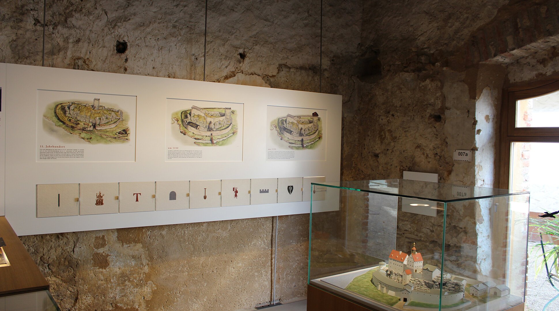 Ausstellungsraum mit historischen Burgzeichnungen und einem Modell in einer Glasvitrine.