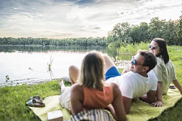 Drei Personen liegen auf einer Decke am Seeufer und schauen auf das Wasser.