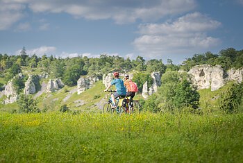 Radfahrer bei den 12 Apostel Solnhofen Zwei Personen fahren mit Fahrrädern auf einem grasbewachsenen Feld vor einer Felslandschaft.