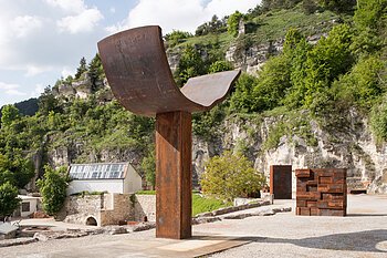 Große Metallskulptur in einer Landschaft mit Felsen und Bäumen im Hintergrund.