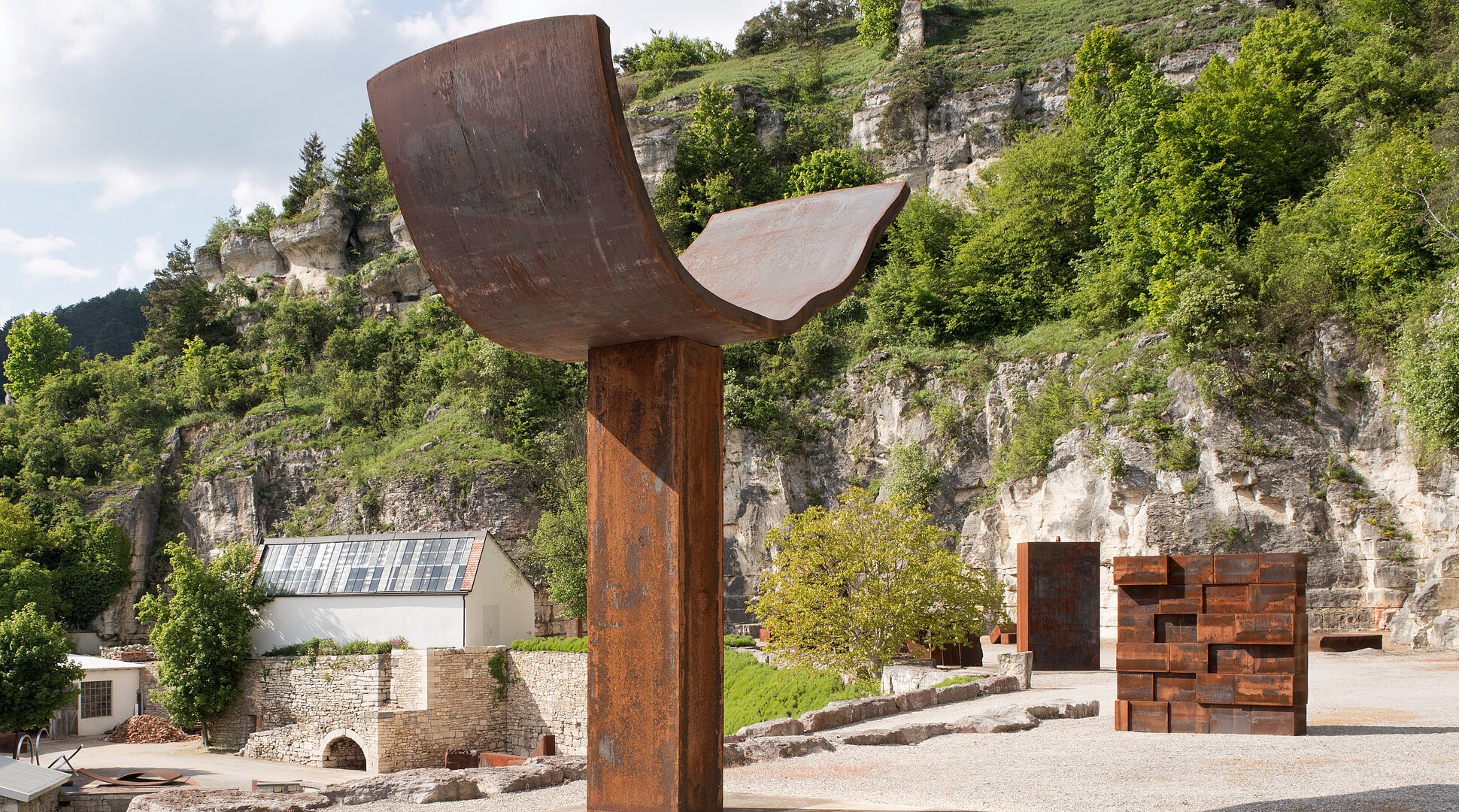 Große Metallskulptur in einer Landschaft mit Felsen und Bäumen im Hintergrund.
