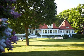 Wittelsbacher Golfclub Außenansicht Ein großes Haus mit roten Dächern und weißen Fensterrahmen, umgeben von Bäumen und Rasenfläche.