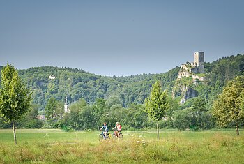 Zwei Radfahrer auf einem Weg vor grüner Wiese, Bäumen und bewaldetem Hügel mit Burgruine im Hintergrund.