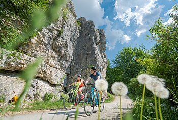 Zwei Radfahrer fahren auf einem Weg neben Felsen und grünen Bäumen bei blauem Himmel mit Wolken.