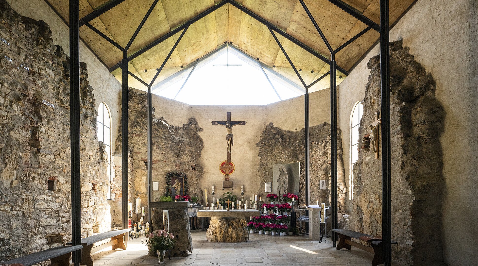 Innenraum einer Kapelle mit Altar, Kreuz, Kerzen und Blumen, umgeben von alten Steinmauern und Holzdecke