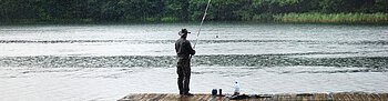 Person steht auf Holzsteg und angelt an einem See mit bewaldetem Ufer im Hintergrund.