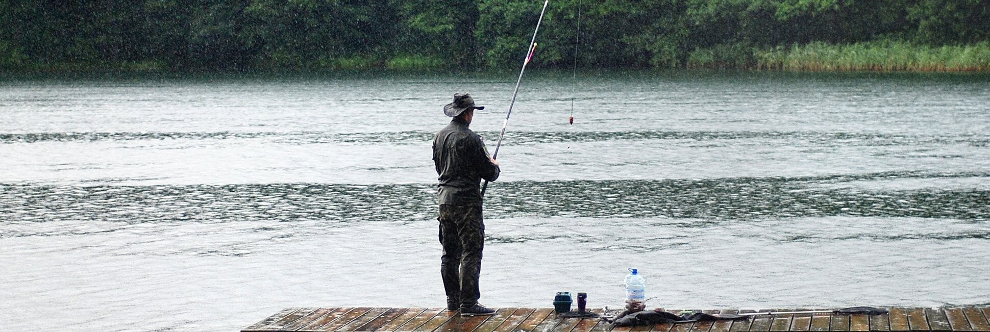 Person steht auf Holzsteg und angelt an einem See mit bewaldetem Ufer im Hintergrund.