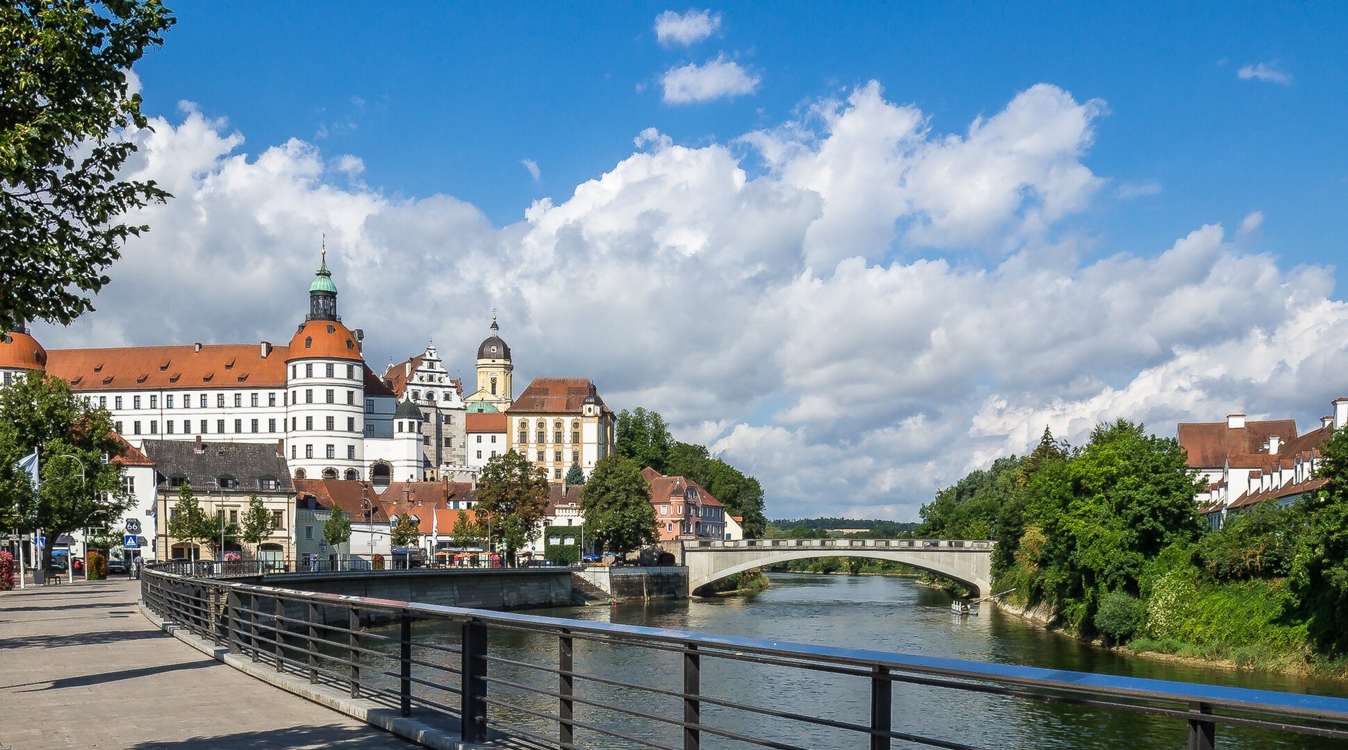 Fluss mit Brücke, rechts Bäume und Häuser, links Blumen, Baum und Schloss im Hintergrund bei blauem Himmel.