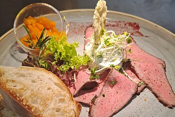 Teller mit Roastbeef, Salat, Brot, Frischkäse mit Sprossen und einem kleinen Glas mit orangener Sauce.