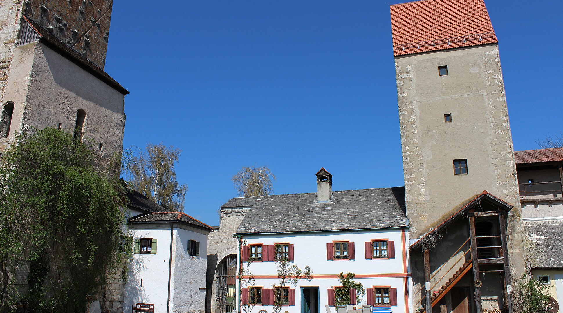 Bauernhof mit weißen Gebäuden, zwei Türmen und Gartenmöbeln unter blauem Himmel.