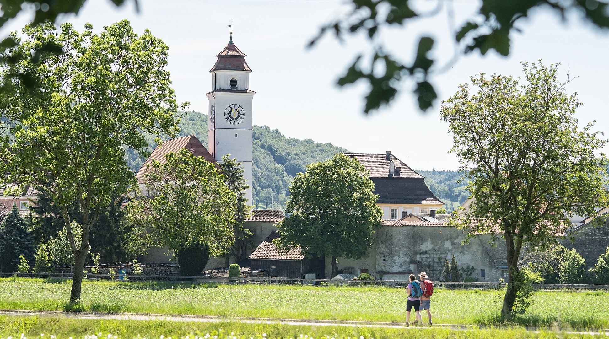 Zwei Personen gehen auf einem Weg durch eine grüne Wiese, im Hintergrund ein Kirchturm und Bäume.