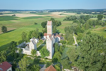 Luftaufnahme einer mittelalterlichen Burg mit drei Türmen umgeben von Feldern und Bäumen an einem sonnigen Tag.