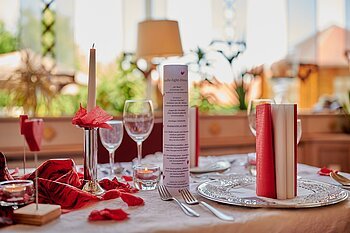 Romantisch gedeckter Tisch mit Kerze, Weingläsern und roter Dekoration bei Tageslicht.