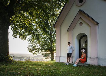 Mann lehnt an Hauswand, Frau sitzt in Türbogen, große Bäume, Dorf im Hintergrund bei Tageslicht.