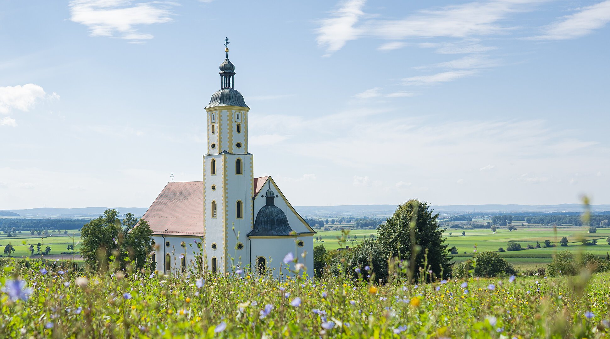 Kirche mit Turm hinter blühendem Feld unter blauem Himmel mit Wolken in ländlicher Umgebung