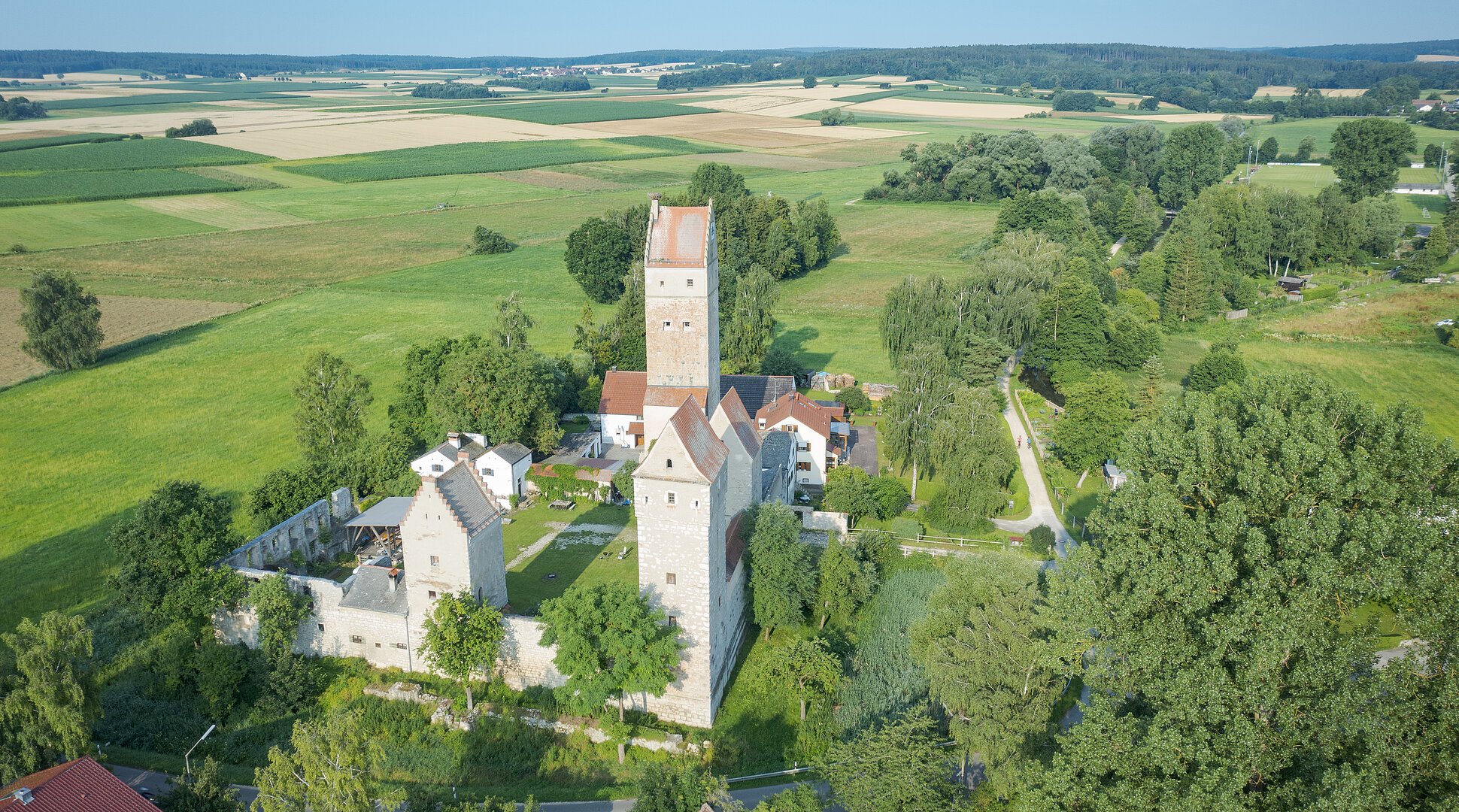 Luftaufnahme einer mittelalterlichen Burg mit drei Türmen umgeben von Feldern und Bäumen an einem sonnigen Tag.