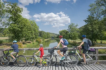 Vier Personen mit Fahrrädern auf einer Holzbrücke mit Blick auf einen Fluss und grüne Landschaft.