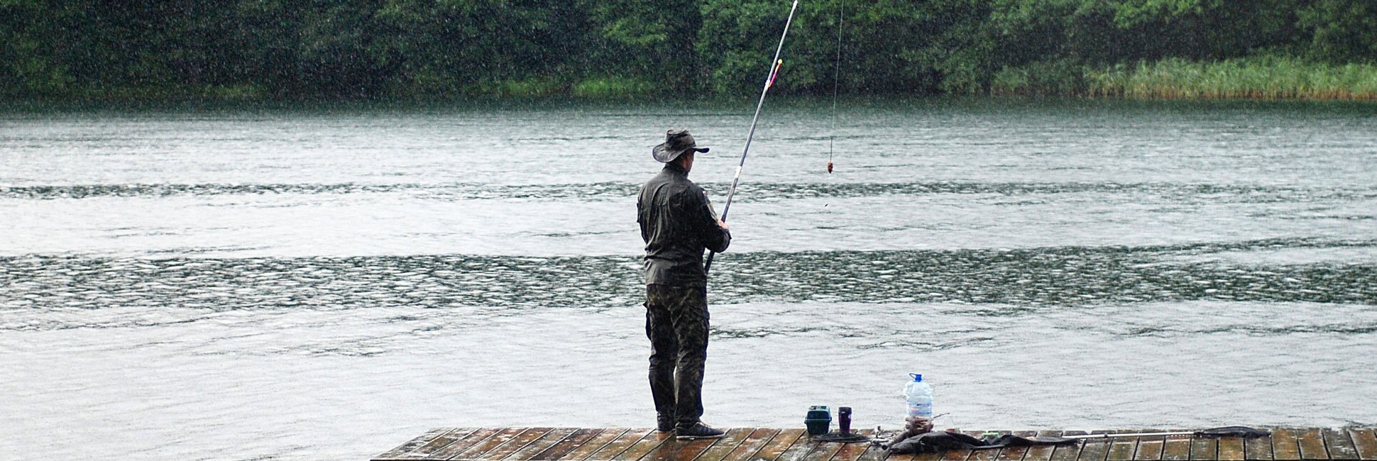 Person steht auf Holzsteg und angelt an einem See mit bewaldetem Ufer im Hintergrund.