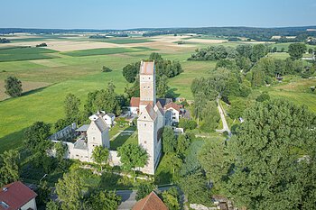 Luftaufnahme einer mittelalterlichen Burg mit drei Türmen umgeben von Feldern und Bäumen an einem sonnigen Tag.