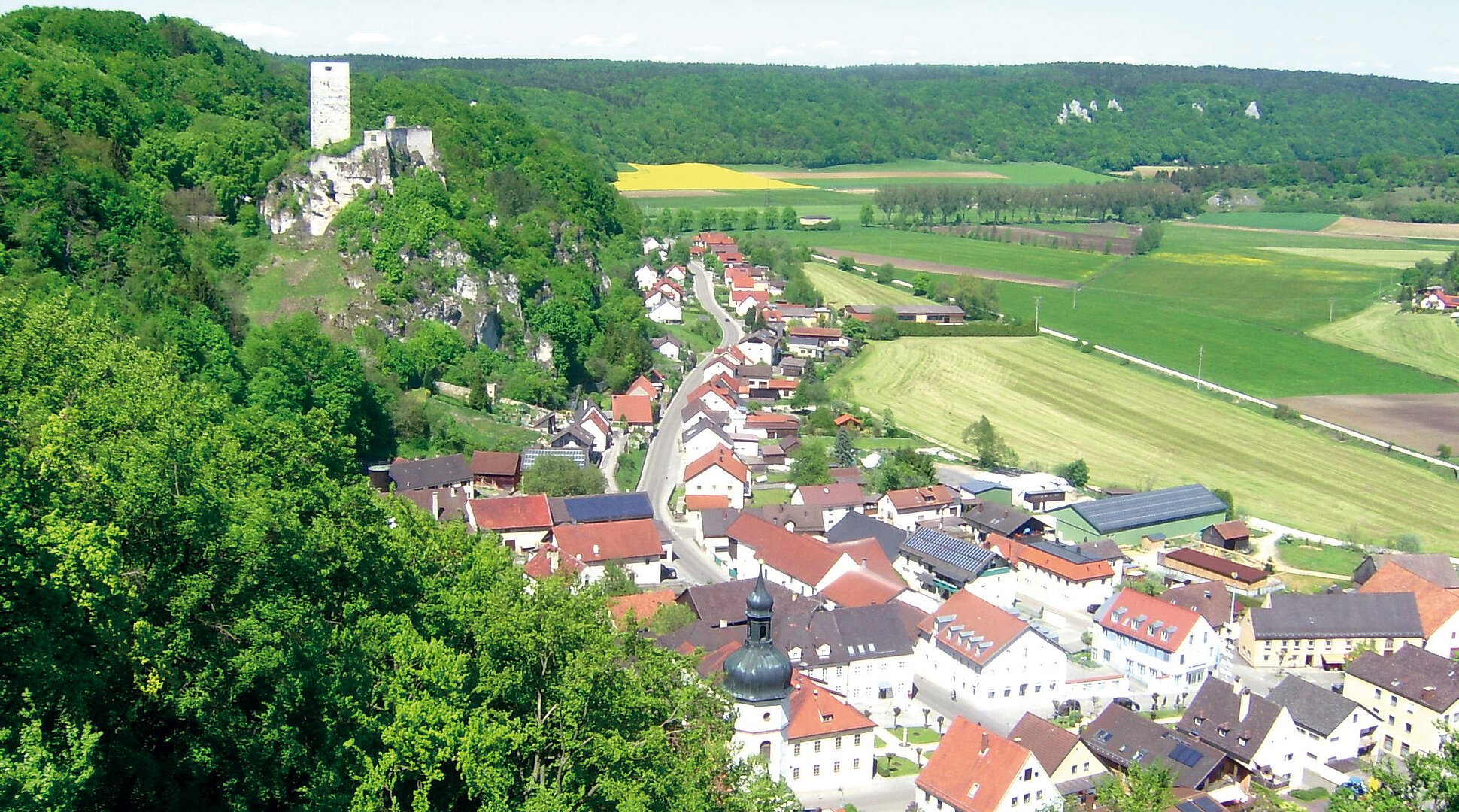 Blick auf ein Dorf mit Kirche und Burgruine auf bewaldetem Hügel, umgeben von Feldern und Wiesen.