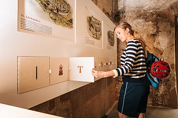 Frau in gestreiftem Pullover öffnet Klappe an Ausstellungstafel mit historischen Texten und Bildern.