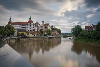 Schloss Neuburg Fluss mit Brücke und historischen Gebäuden im Hintergrund, bewölkter Himmel.