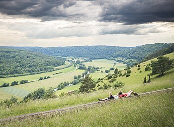 Zwei Wanderinnen liegen auf einer langen Holzbank und schauen ins weitläufige grüne Altmühltal.