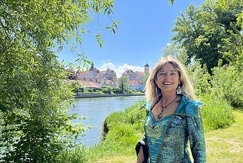 Die Stadtführerin steht im Kostüm der Danubia am grasigen Donauufer. Sie trägt ein blau und grün schimmerndes Kleid. Im Hintergrund fließt die Donau. Das gegenüberliegende Ufer ist zu erkennen, man kann die Stadt Neuburg erkennen. Die Sonne scheint.