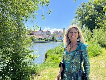Die Stadtführerin steht im Kostüm der Danubia am grasigen Donauufer. Sie trägt ein blau und grün schimmerndes Kleid. Im Hintergrund fließt die Donau. Das gegenüberliegende Ufer ist zu erkennen, man kann die Stadt Neuburg erkennen. Die Sonne scheint.