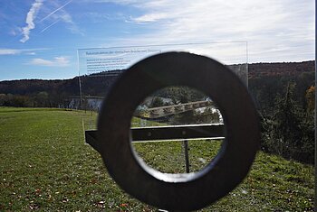 Rennertshofen Aussichtspunkt Antoniberg Infotafel Blick durch ein rundes Metallrohr auf eine Infotafel mit Brückenrekonstruktion vor einer Wiese und Wald im Hintergrund.