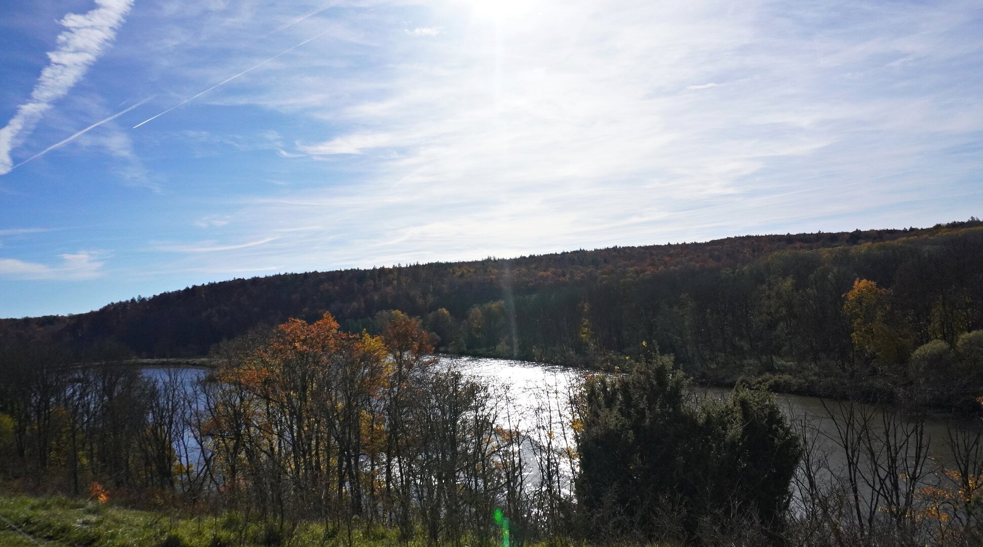 Fluss mit Bäumen am Ufer und bewaldetem Hügel unter blauem Himmel mit Sonne und Wolken.
