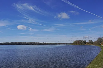 Uferweg entlang eines großen Gewässers unter blauem Himmel mit wenigen Wolken und Baumreihe am Horizont.