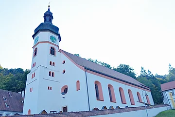 Kirchturm mit Uhr an weißem Kirchengebäude vor bewaldetem Hügel unter hellem Himmel