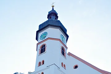 Blick durch ein Metalltor auf eine weiße Kirche mit Turmuhr und Treppe, im Hintergrund Bäume.
