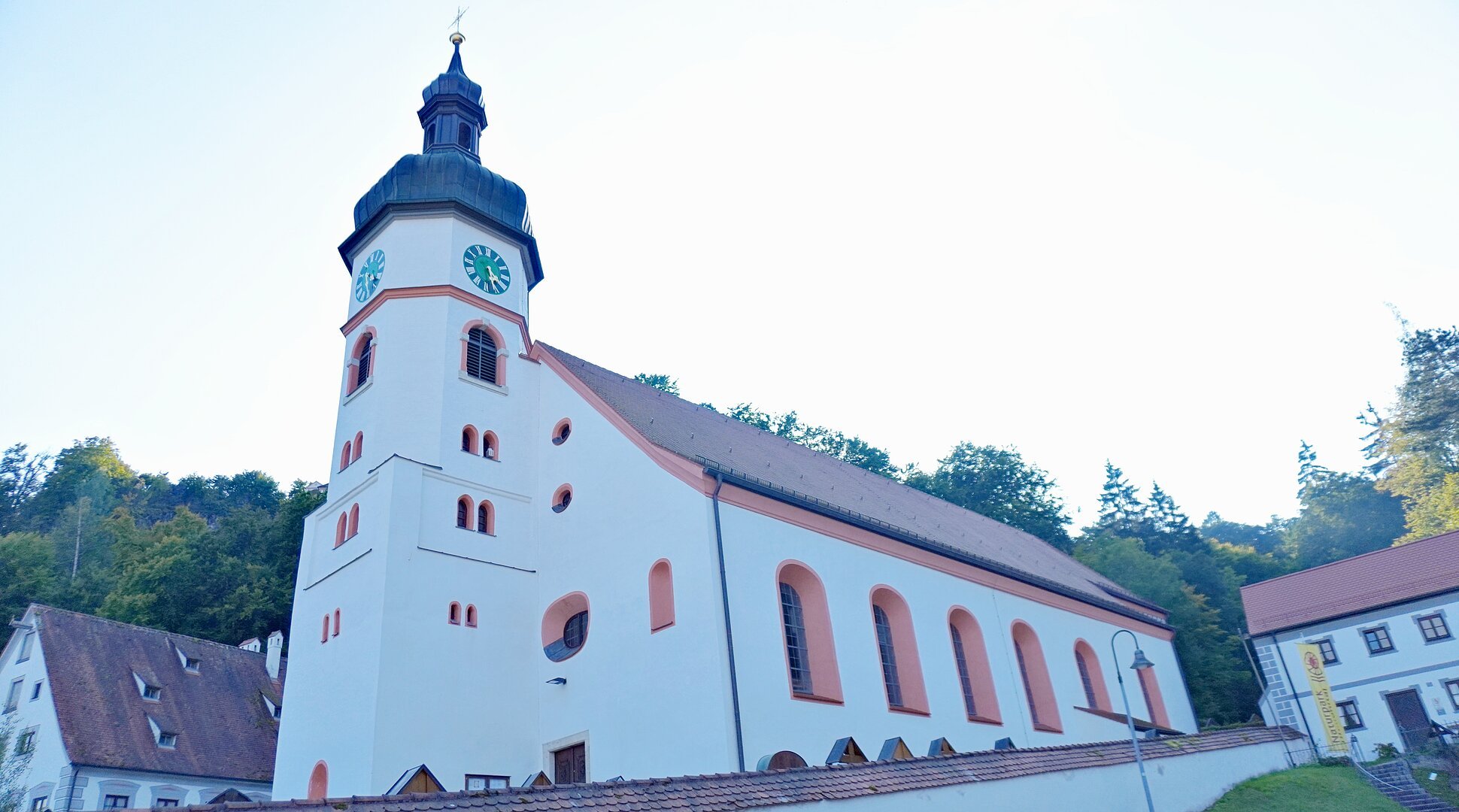Kirchturm mit Uhr an weißem Kirchengebäude vor bewaldetem Hügel unter hellem Himmel