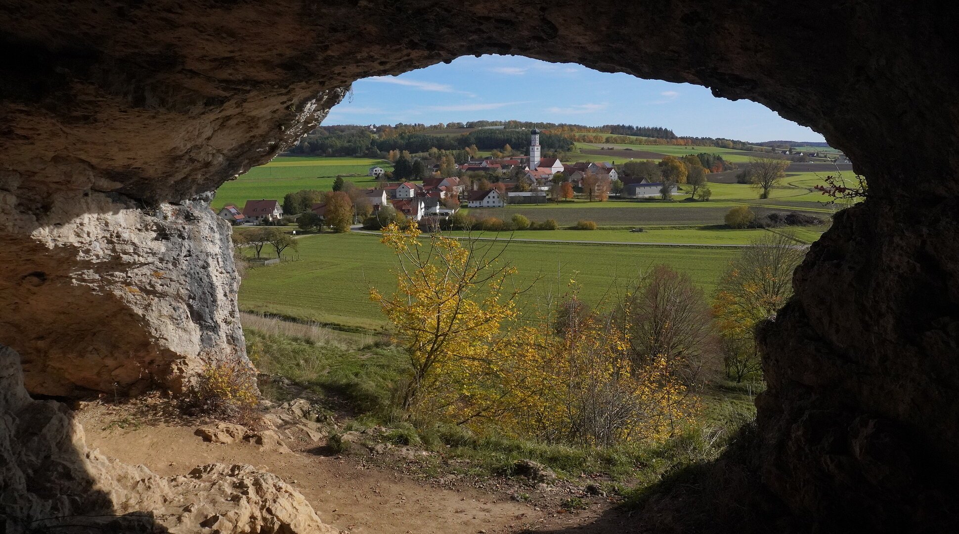 Blick aus einer Höhle auf ein Dorf mit Kirche, Feldern und Bäumen im Herbst bei klarem Himmel.