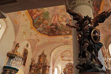 Innenraum einer Kirche mit bemaltem Gewölbe und Statue eines Engels mit Schild und Schwert an einer Säule.