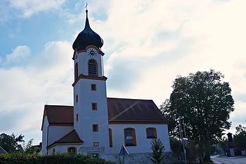 Kirchturm mit Zwiebelhaube und Uhr an weißem Kirchengebäude bei bewölktem Himmel und Bäumen im Hintergrund.