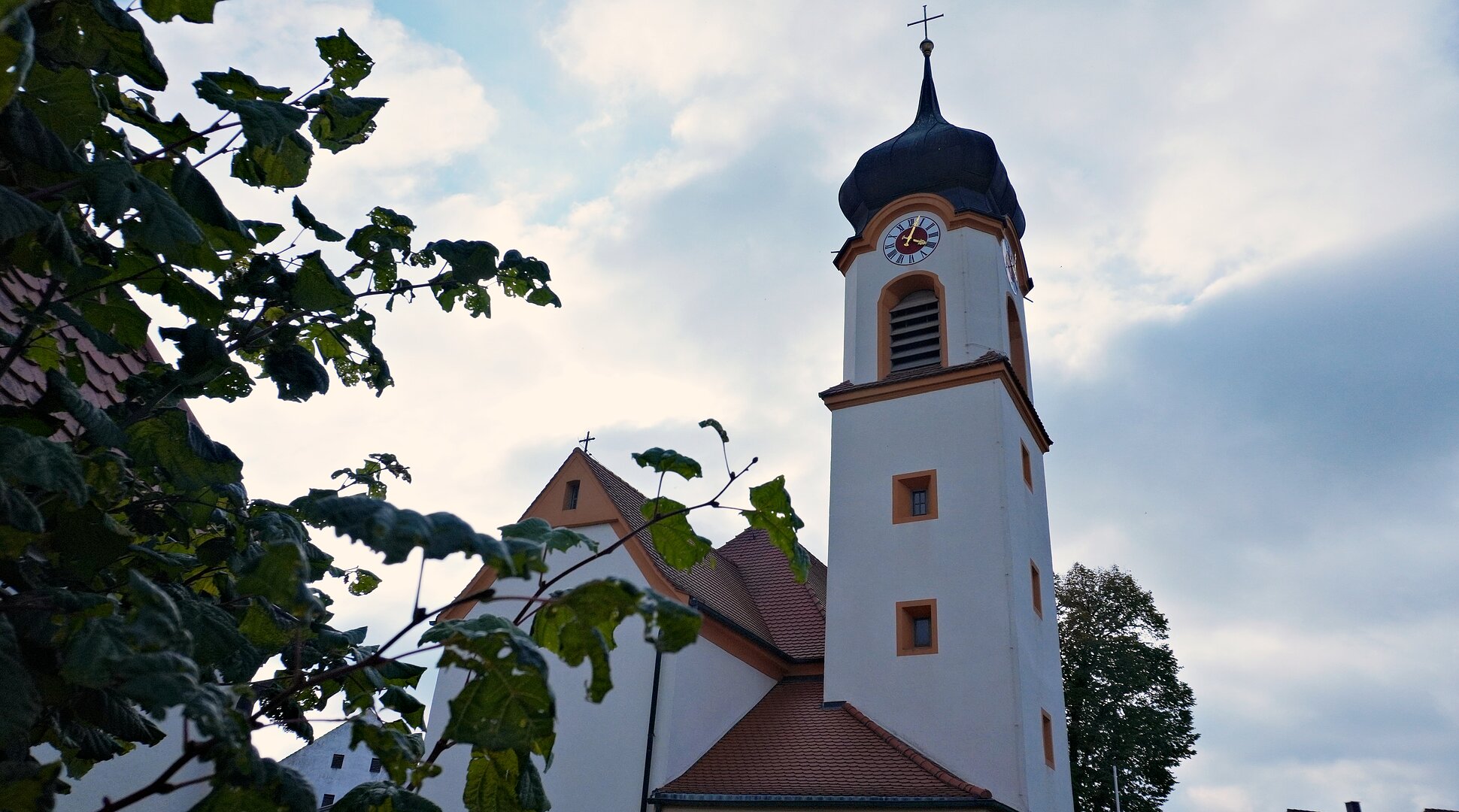 Kirchturm mit Uhr und Kreuz, teilweise von Blättern vor bewölktem Himmel verdeckt.