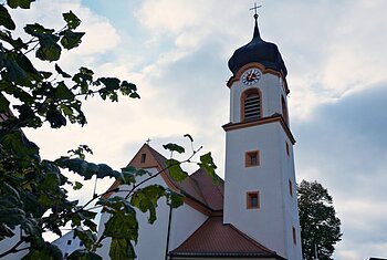 Kirchturm mit Uhr und Kreuz, teilweise von Blättern vor bewölktem Himmel verdeckt.