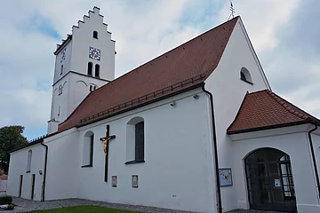 Weiße Kirche mit rotem Dach, Uhrturm und großem Kreuz an der Außenwand bei bewölktem Himmel.
