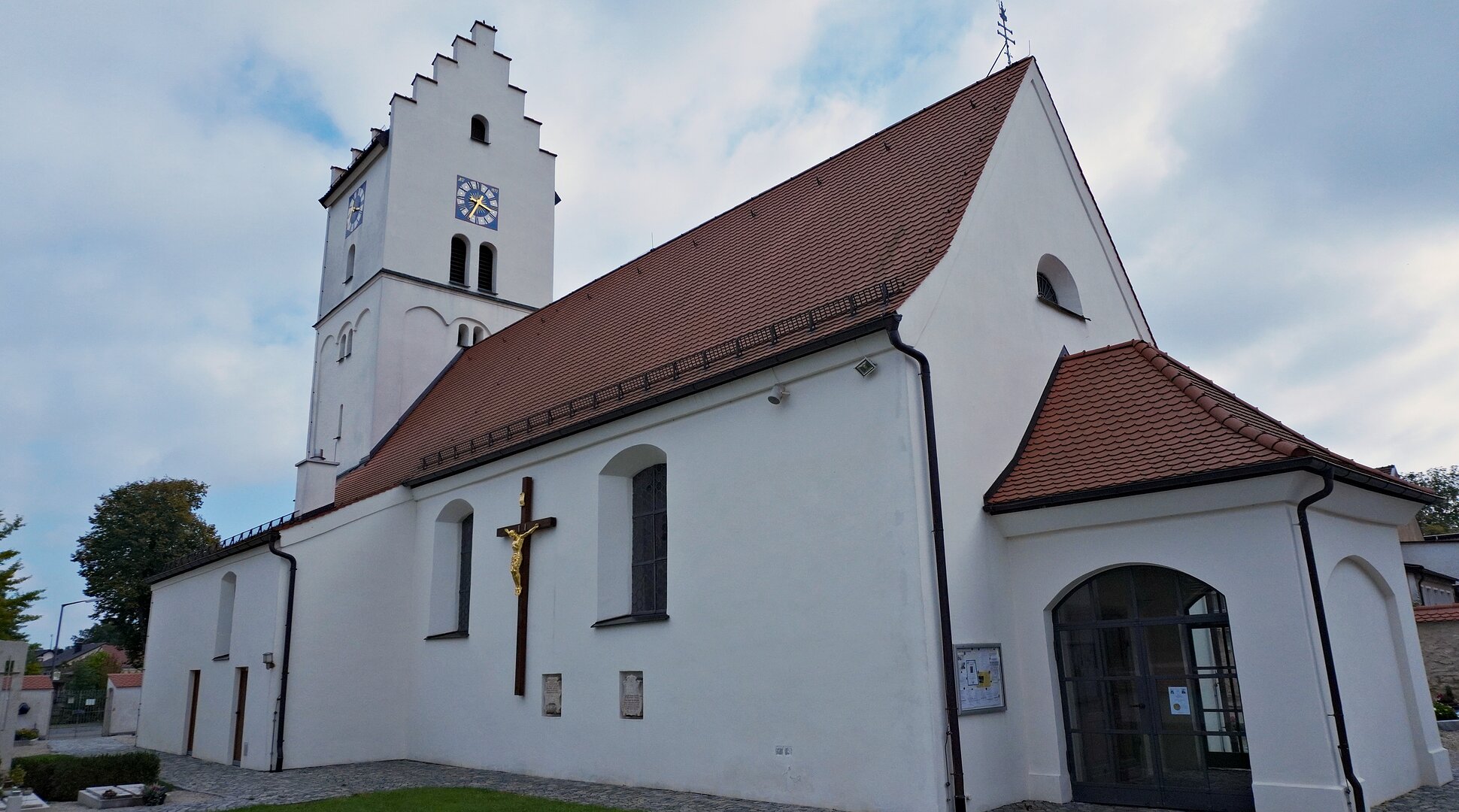 Weiße Kirche mit rotem Dach, Uhrturm und großem Kreuz an der Außenwand bei bewölktem Himmel.